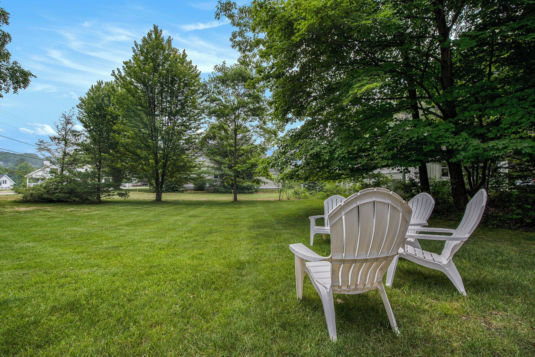 Un espace extérieur verdoyant, avec des chaises Adirondack blanches et une table, entouré d’arbres et avec une maison en arrière-plan.