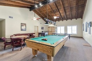 Une table de billard dans une pièce dotée d’un plafond haut et de poutres en bois, un coin repas avec des chaises et une table, et un ventilateur de plafond.