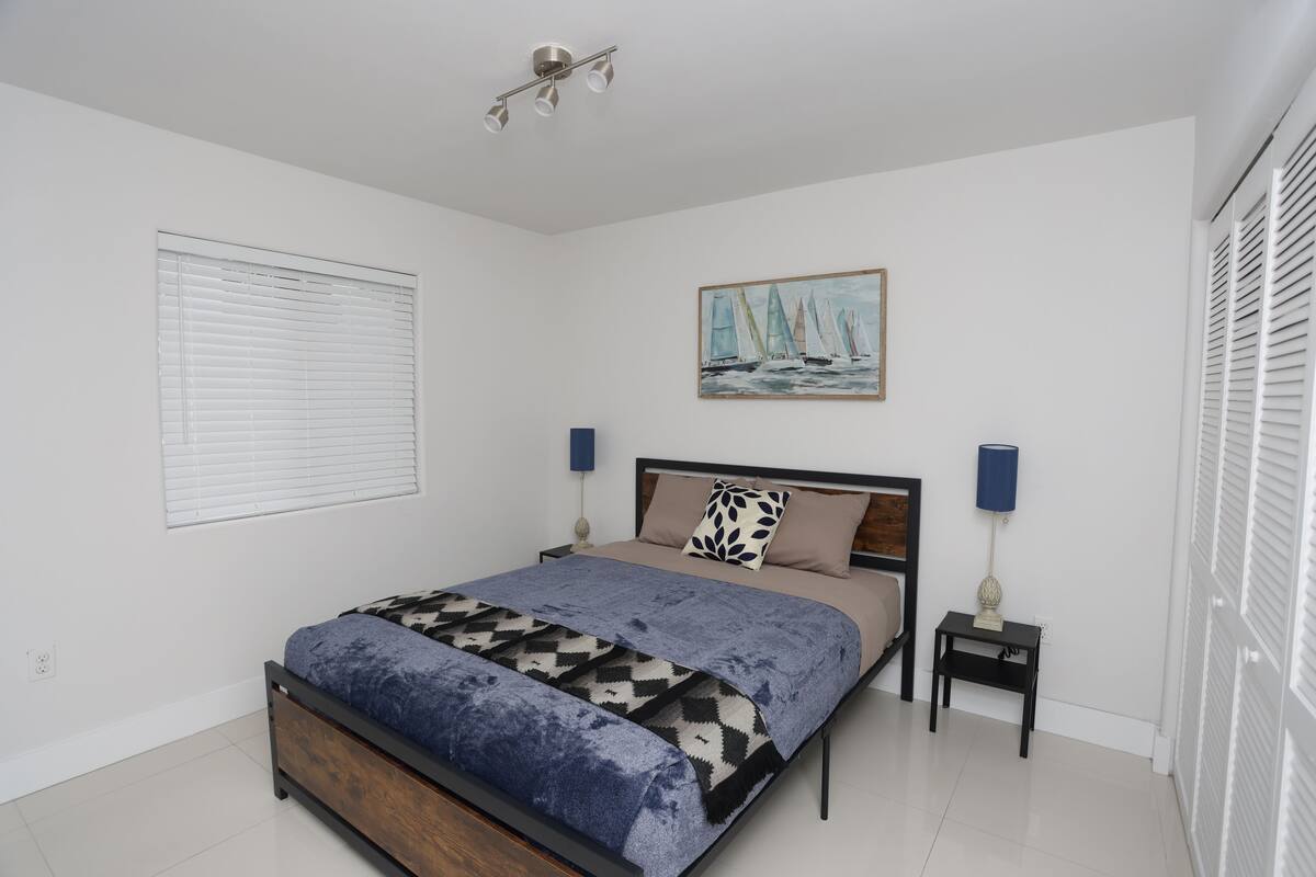 A bedroom with a bed, a painting, two bedside tables, and a window with blinds.