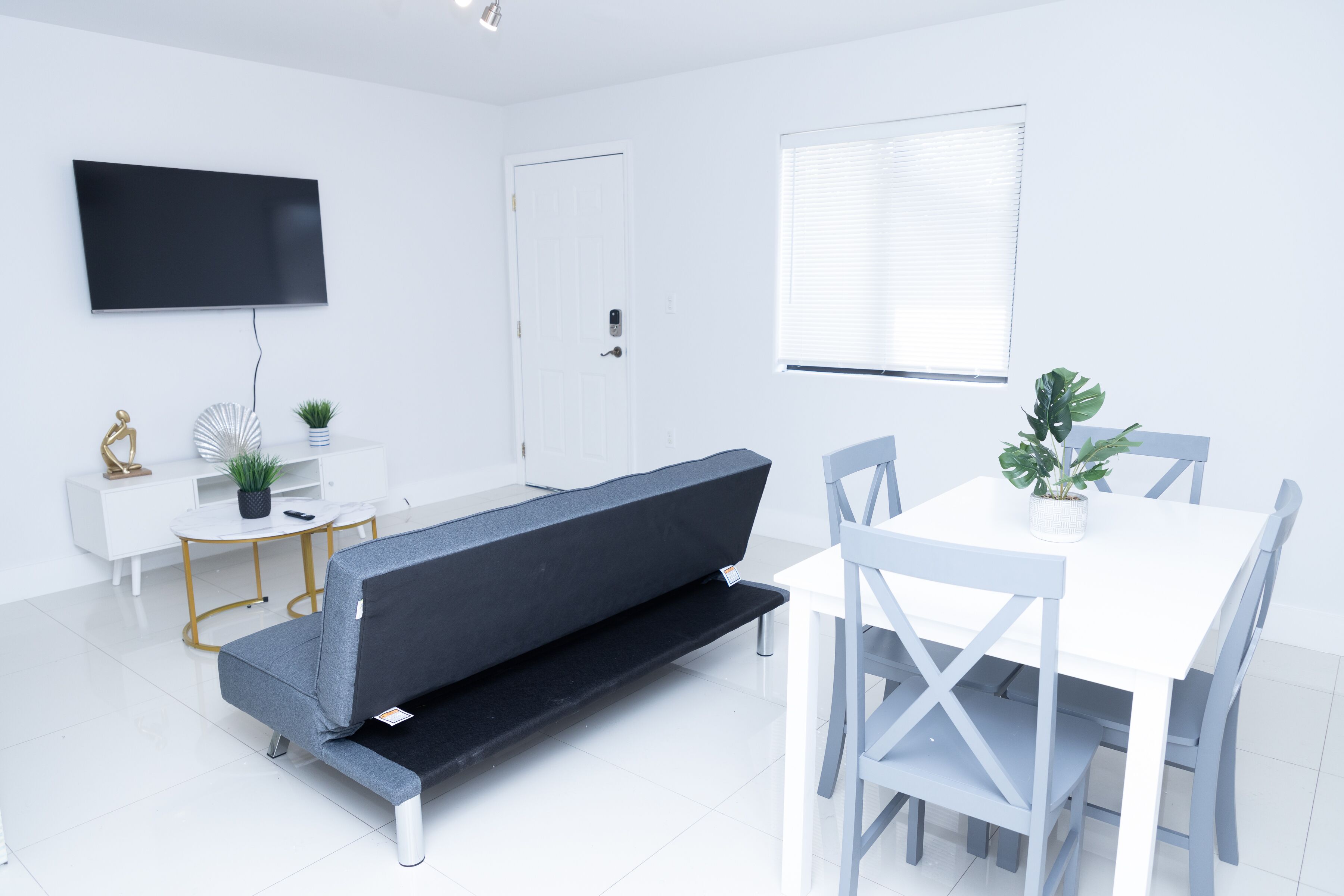 A modern, minimalist room with a white sofa, a dining table with chairs, a flat-screen TV mounted on the wall, and a window with blinds.