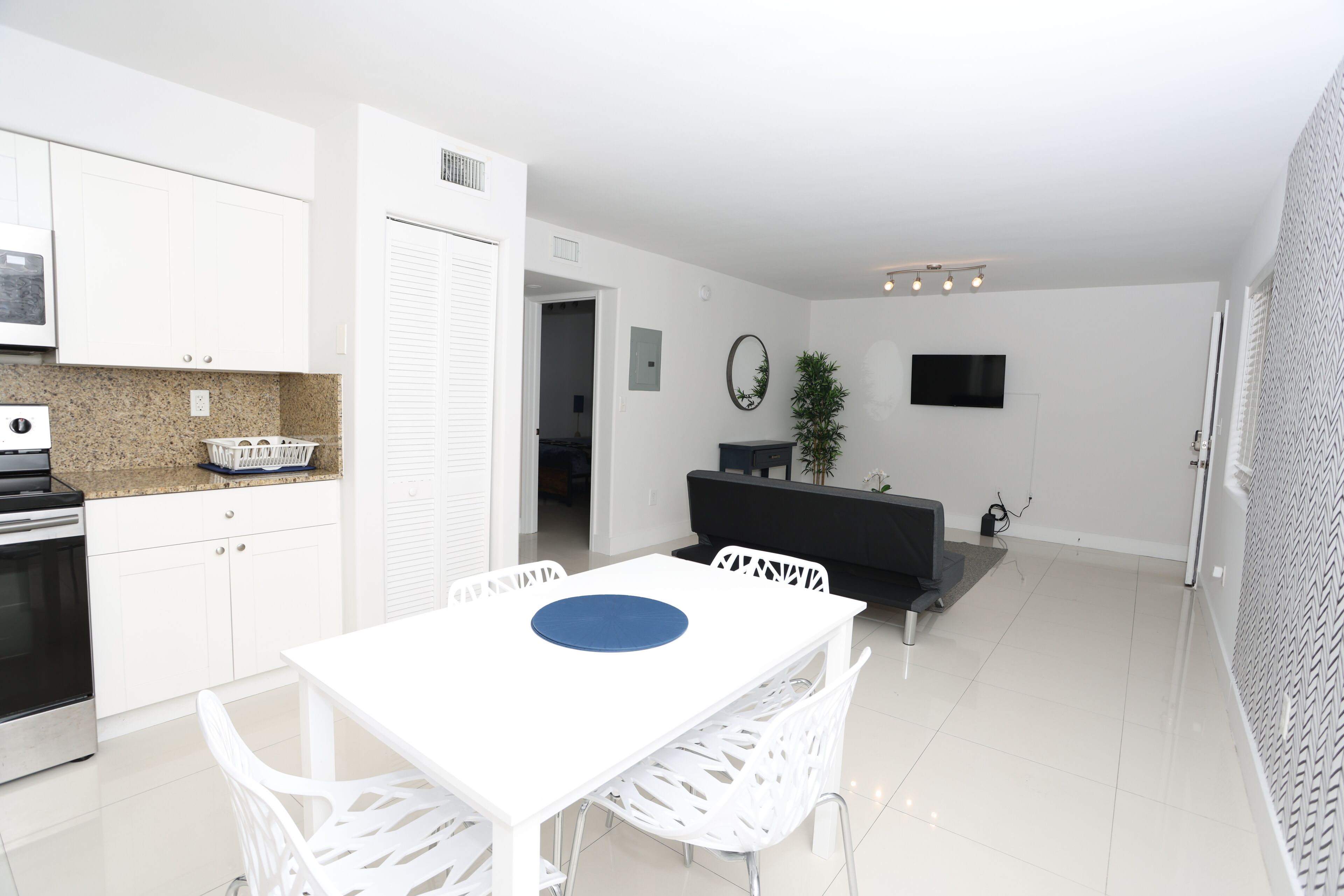 A modern kitchen with white cabinets, a granite countertop, and a built-in oven. There is a dining area with a white table and chairs, and a black sofa in the background.