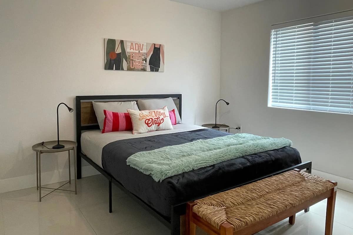 A bedroom with a bed, bedside tables, a bench, a window with blinds, and a wall-mounted artwork.