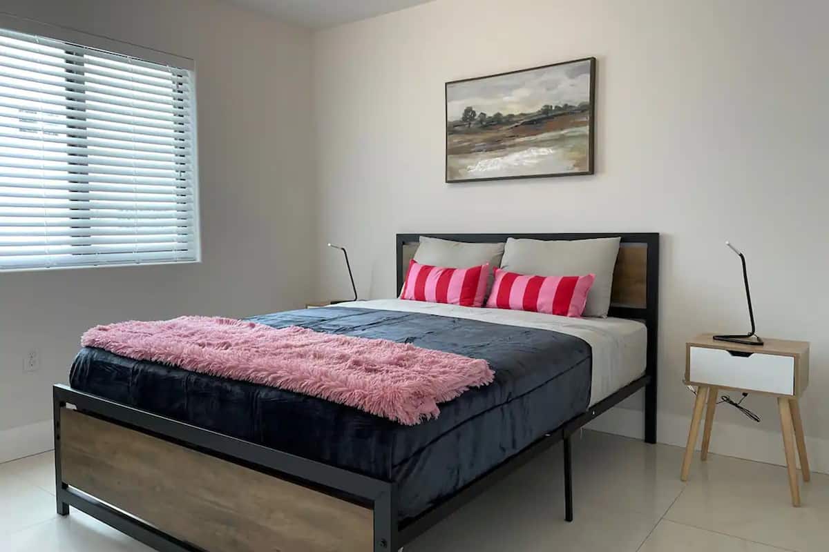 A bedroom with a bed, a bedside table, a window with blinds, and a painting on the wall.