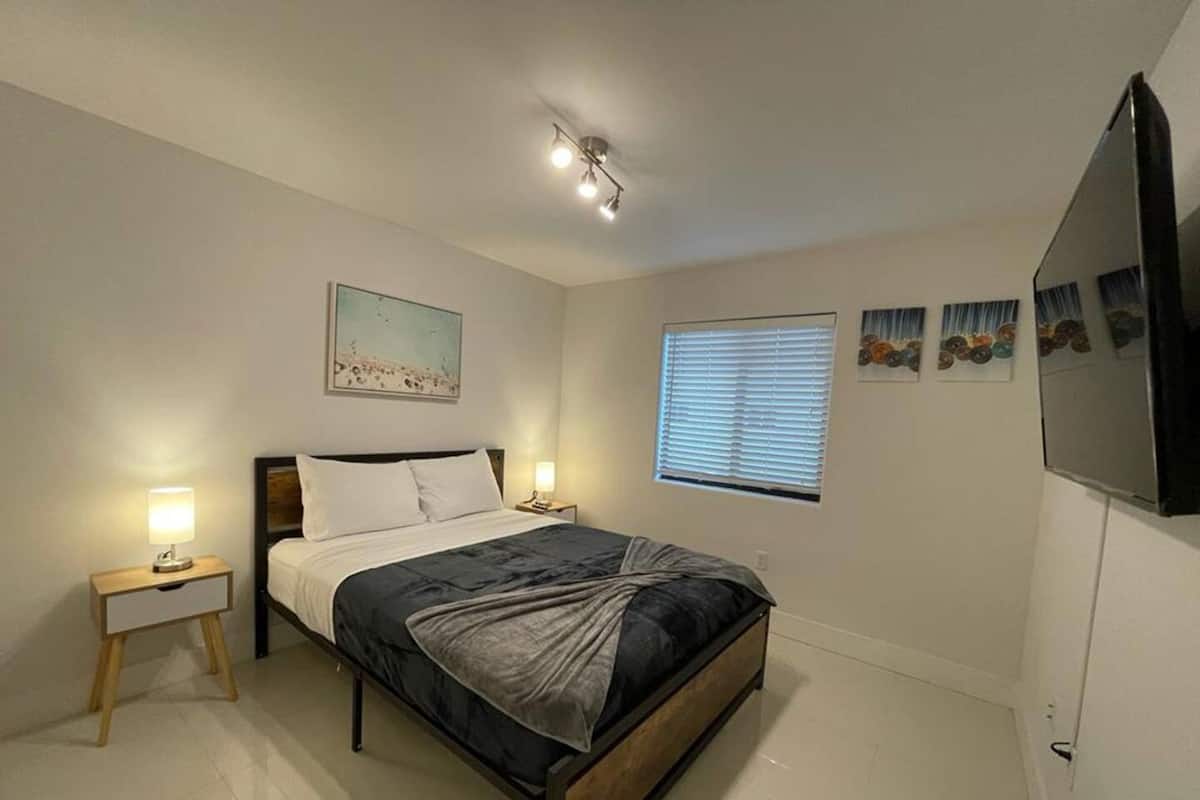 A bedroom with a bed, bedside lamps, a ceiling light, a window with blinds, and wall art.
