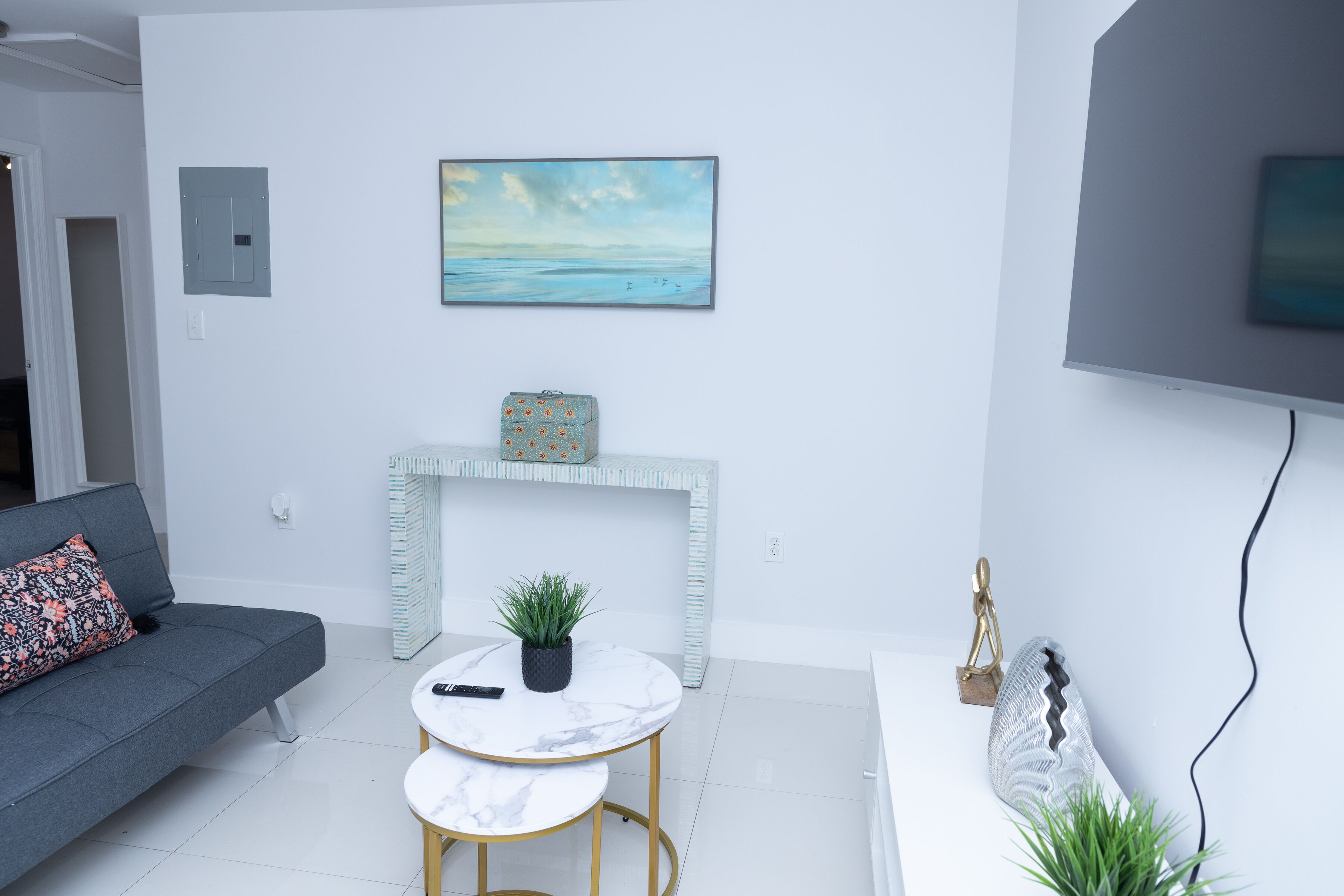 A modern living room with a grey sofa, a white coffee table, a small round table with a plant, a white fireplace, and a painting on the wall.