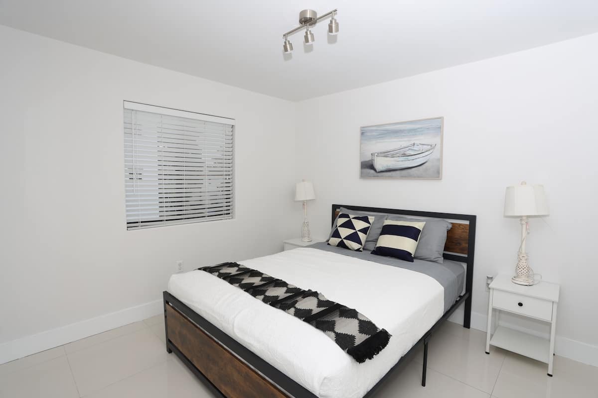 A bedroom with a bed, bedside tables, a window with blinds, a painting, and a ceiling light fixture.