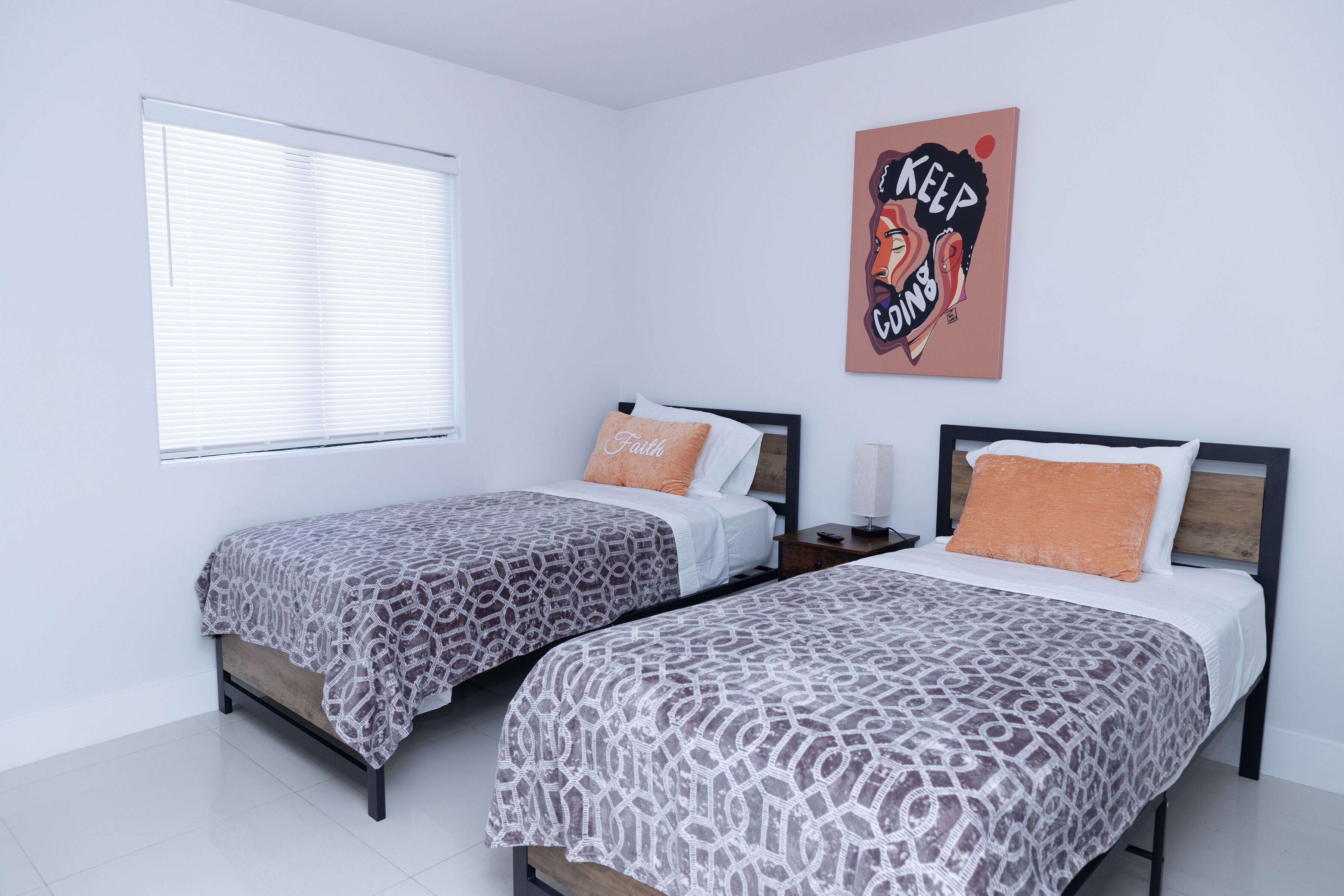 A bedroom with two beds, white walls, a window with blinds, and a wall art piece.