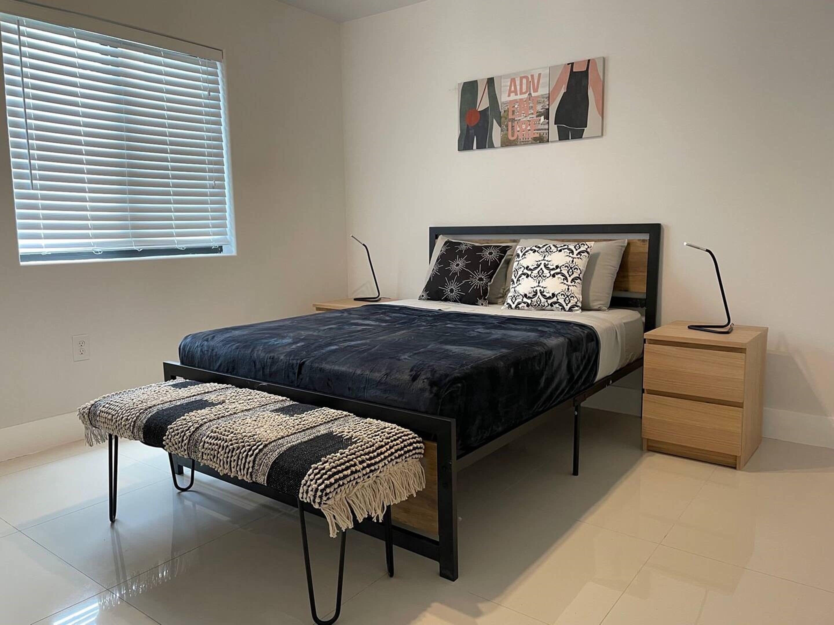 Une chambre moderne avec un lit, des tables de chevet, un banc et une fenêtre avec des stores.