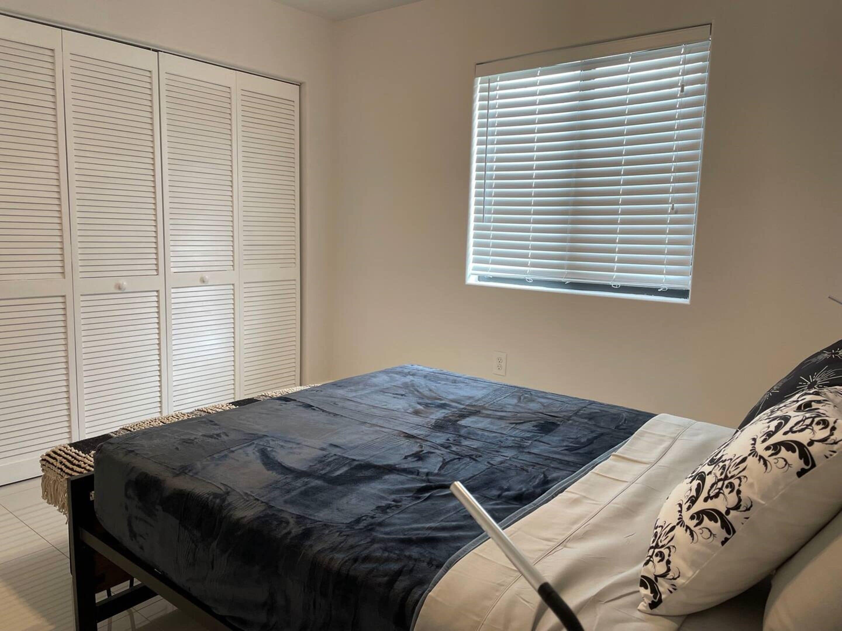 Quarto com cama, janela com persianas e guarda-roupa com portas venezianas.