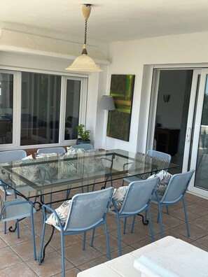 Une table à manger avec un plateau en verre, entourée de chaises bleues, un ventilateur de plafond et une grande fenêtre donnant sur un beau paysage.