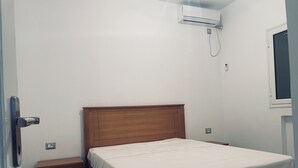 Um quarto com uma cama de madeira, uma mesa de cabeceira, um ar condicionado fixado na parede e uma janela.
