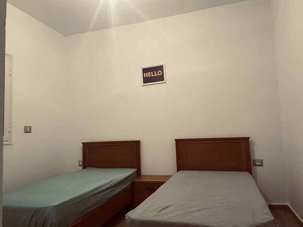 A small, simple room with two single beds, a nightstand, and a 'HELLO' sign on the wall.