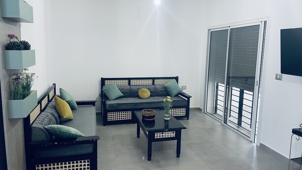 Uma sala de estar moderna com um sofá cinzento, uma poltrona preta e uma mesa de centro. Há uma janela grande com persianas, uma luminária no teto e uma TV fixada na parede.
