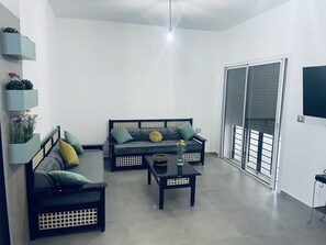A modern living room with a grey sofa, a black armchair, and a coffee table. There is a large window with blinds, a ceiling light, and a wall-mounted TV.