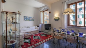 Sala de estar com sofá, televisão, mesa de vidro, cadeiras e um tapete vermelho.