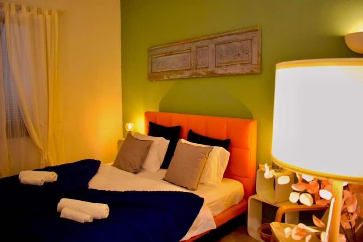Un lit bien fait, avec une couverture bleue et des oreillers blancs, une lampe de chevet et une œuvre d’art accrochée au mur.