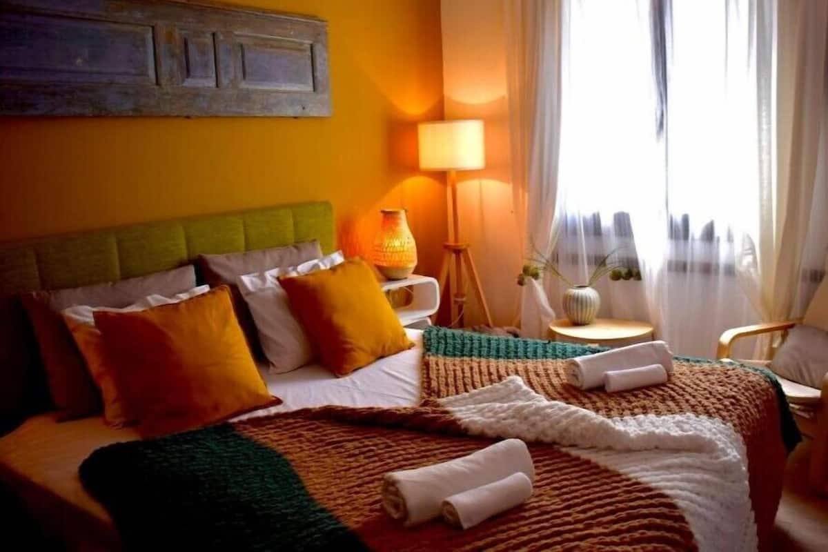 Une chambre d’hôtel avec une tête de lit recouverte d’un tissu vert, un lit avec une literie blanche et orange, une lampe de chevet et une vue sur la ville à travers des rideaux transparents.