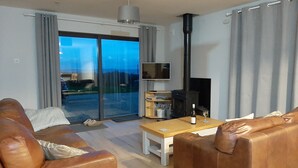 Una sala de estar moderna con sofás de cuero, una mesa de centro de madera, un televisor de pantalla plana, una chimenea y un gran ventanal corredizo que da a un balcón.