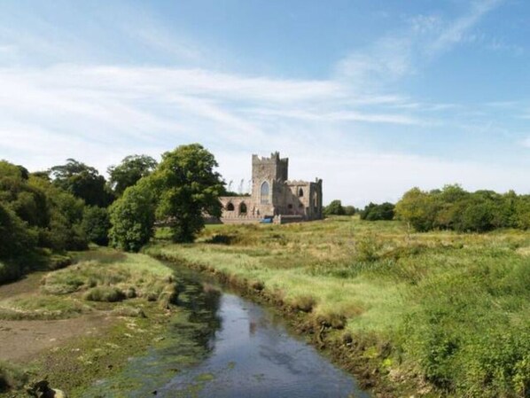 A historic building with a tower, surrounded by greenery and a small stream.
