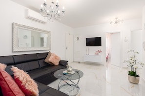 A modern living room with a black sofa, a glass coffee table, a large mirror, a chandelier, and a TV mounted on the wall.