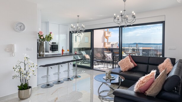 A modern living room with a black sofa, a glass coffee table, and a dining area with a bar. There is a large window offering a view of the city and the sea. - Apartment Summer Breeze Okrug Gornji (Okrug)