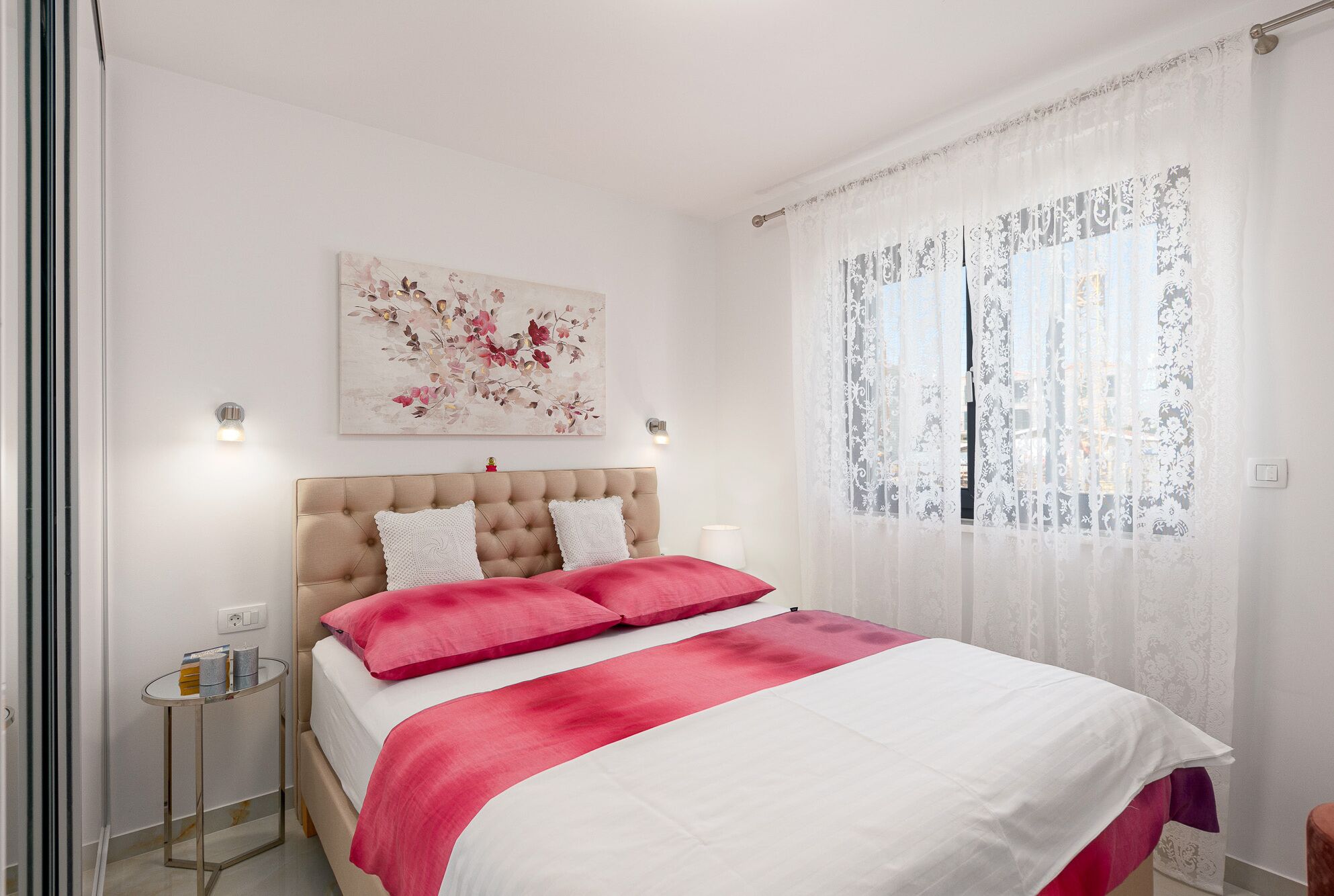 Quarto com cabeceira estofada, cama com roupa de cama rosa e branca, mesa de cabeceira pequena com abajur, luminária na parede e janela com cortinas finas.