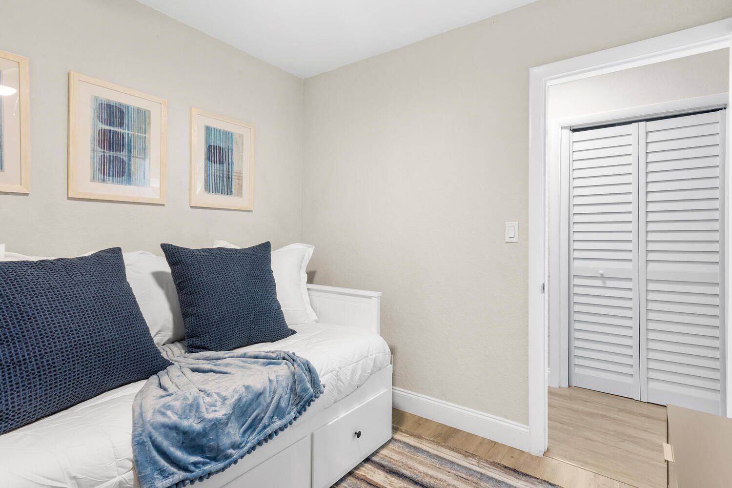 Une chambre à coucher comprenant un lit, une commode, une porte avec des volets à persiennes et trois tableaux encadrés accrochés au mur.