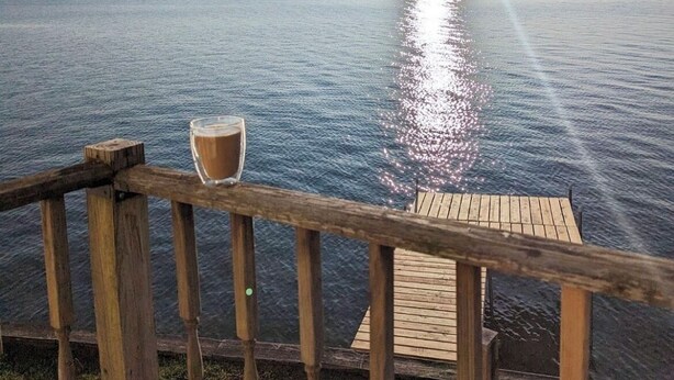 Una terraza de madera con un vaso de bebida y una barandilla de madera, con vistas a un cuerpo de agua tranquilo al atardecer.