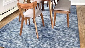 Un comedor con una mesa de madera, seis sillas, un florero con flores y una alfombra azul con textura.