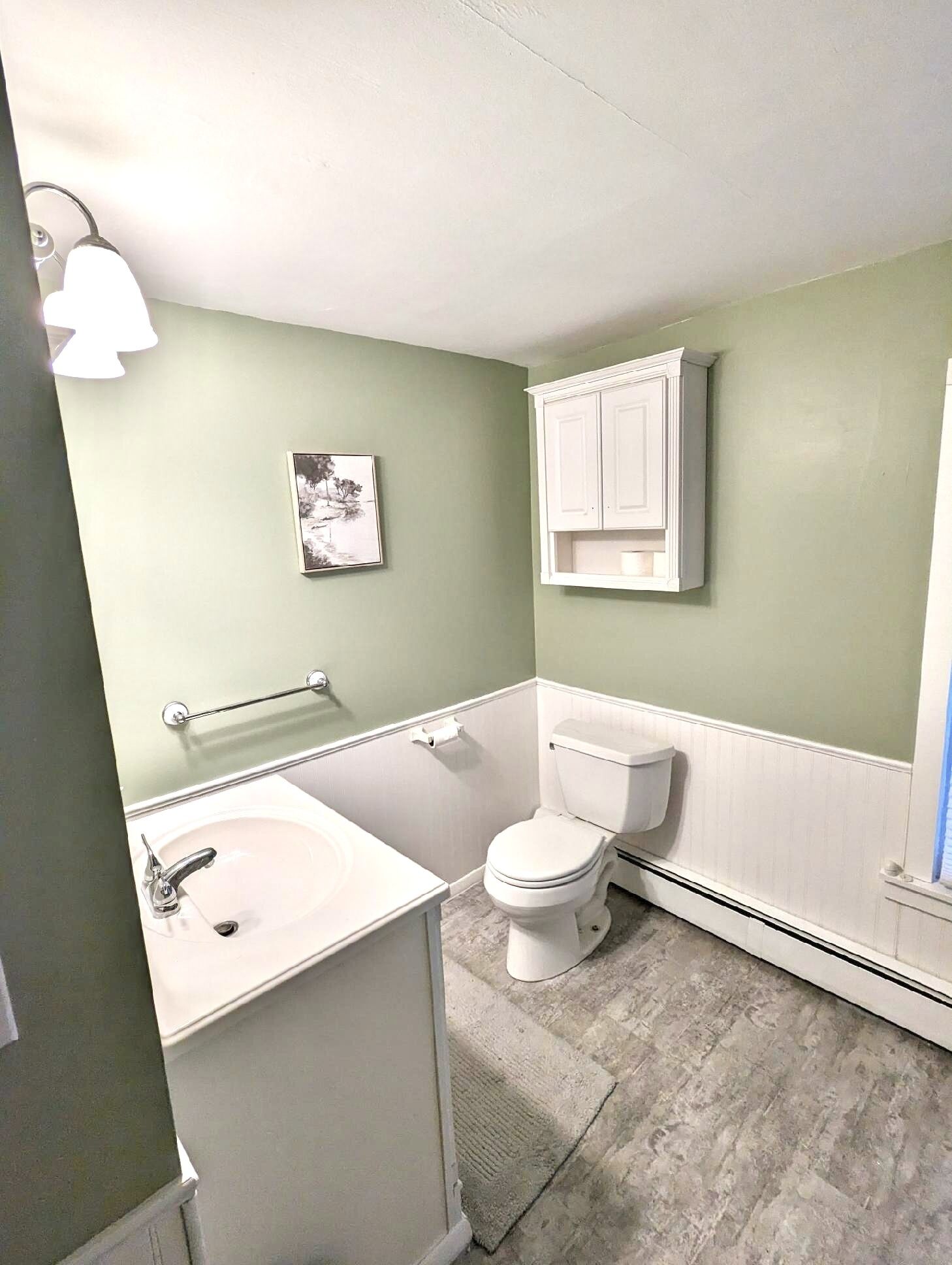 A bathroom with a white sink, toilet, and cabinet, a framed picture on the wall, and a light fixture.