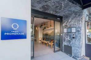 L’entrée d’un hôtel, avec une enseigne indiquant « CRISOL PROMENADE », une porte vitrée et un espace détente offrant une vue sur l’intérieur.