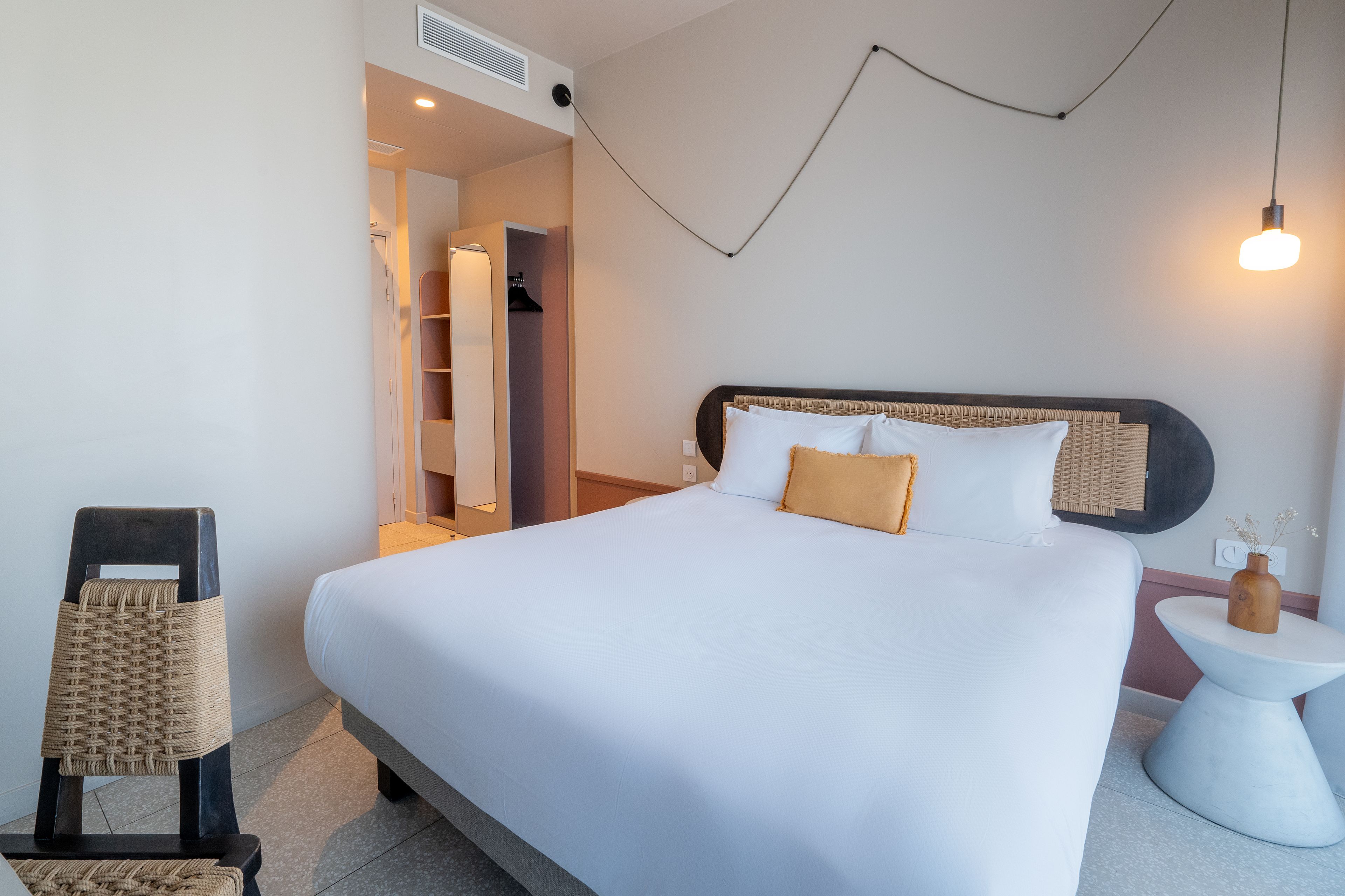 A hotel room with a large bed, a bedside table, a chair, a wardrobe, and a wall-mounted light fixture.