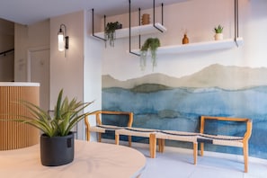 A modern interior with a round table, two wooden benches, a wall-mounted shelf with plants, and a large abstract painting. - Crisol Promenade (Nice)