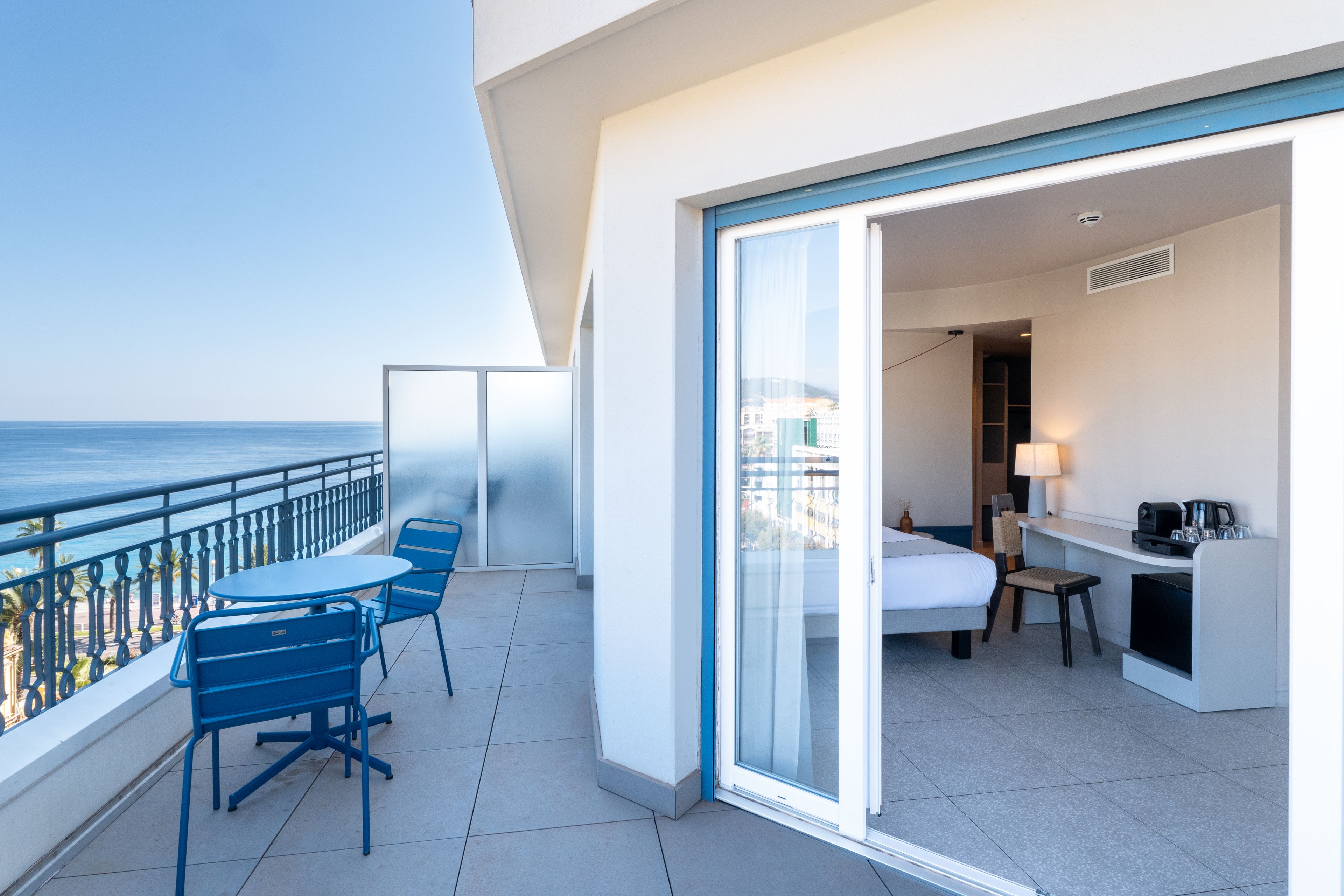 A balcony with a table and chairs, a bed, a desk with a lamp, and a view of the sea.