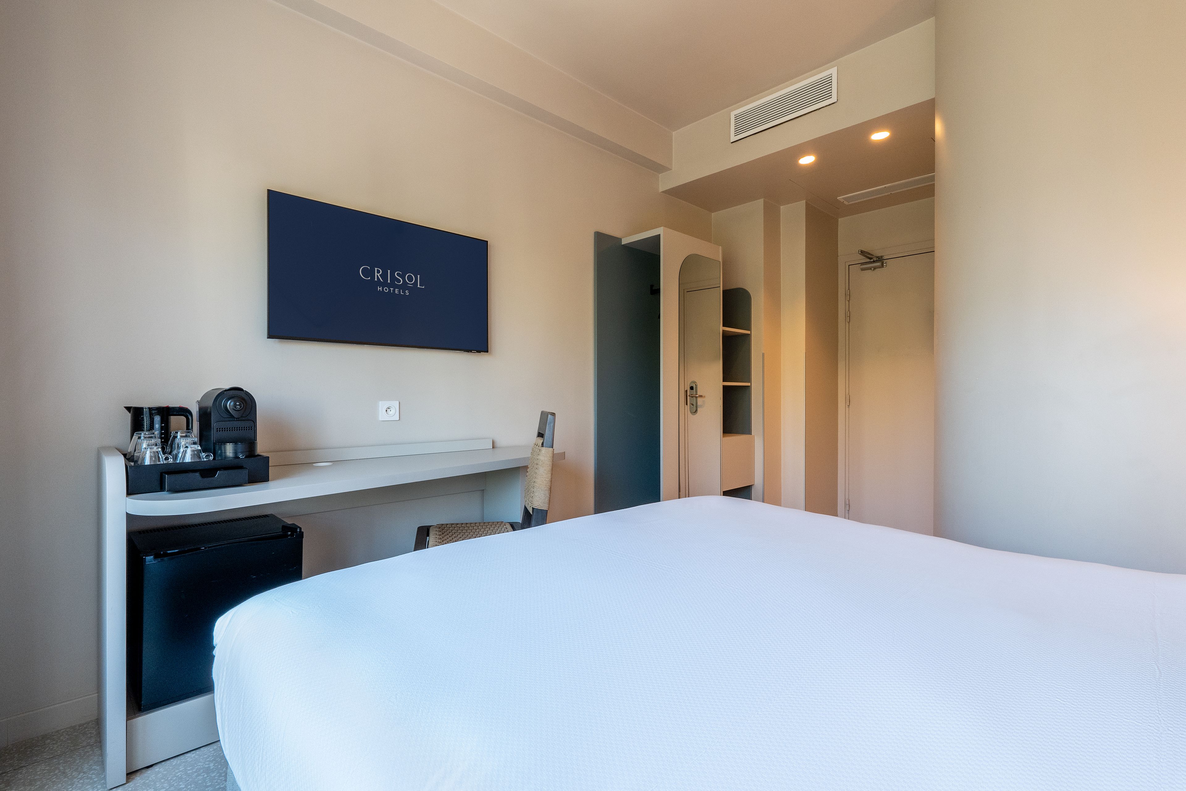 A hotel room with a bed, a desk with a coffee machine, a minibar, and a door leading to a bathroom.