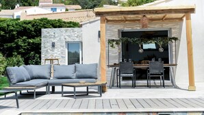 A poolside area with a wooden deck, a gray sofa, a table with chairs, and a pergola with hanging plants. - Splendid villa with swimming pool & sea view (Roquebrune-sur-Argens)