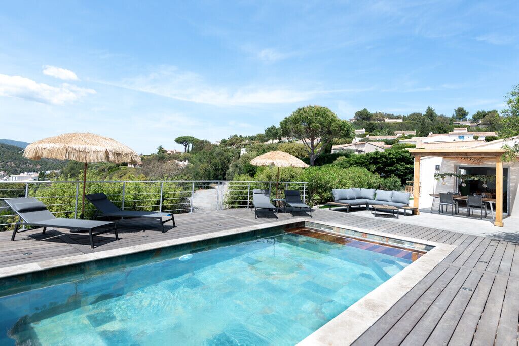 A rooftop pool with lounge chairs and umbrellas, surrounded by greenery and overlooking a hillside.