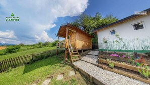 A wooden cabin with a ladder leading to a lofted sleeping area, surrounded by a fenced garden and a white building with a mural. - Blue bungalow Bosnian pyramid glamping (Visoko)