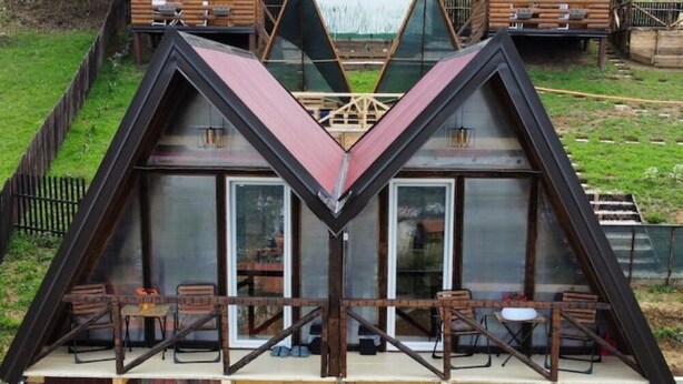 A triangular wooden cabin with a glass facade, outdoor seating, and a small deck. - Red bungalow Bosnian pyramid glamping (Visoko)