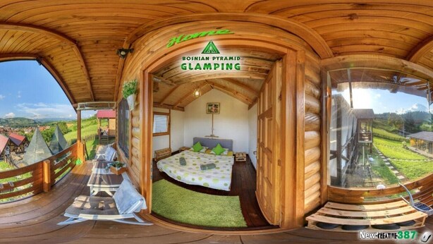 A panoramic view of a wooden log cabin with a bed, a small table, and a window overlooking a scenic landscape. - Green bungalow Bosnian pyramid glamping (Visoko)