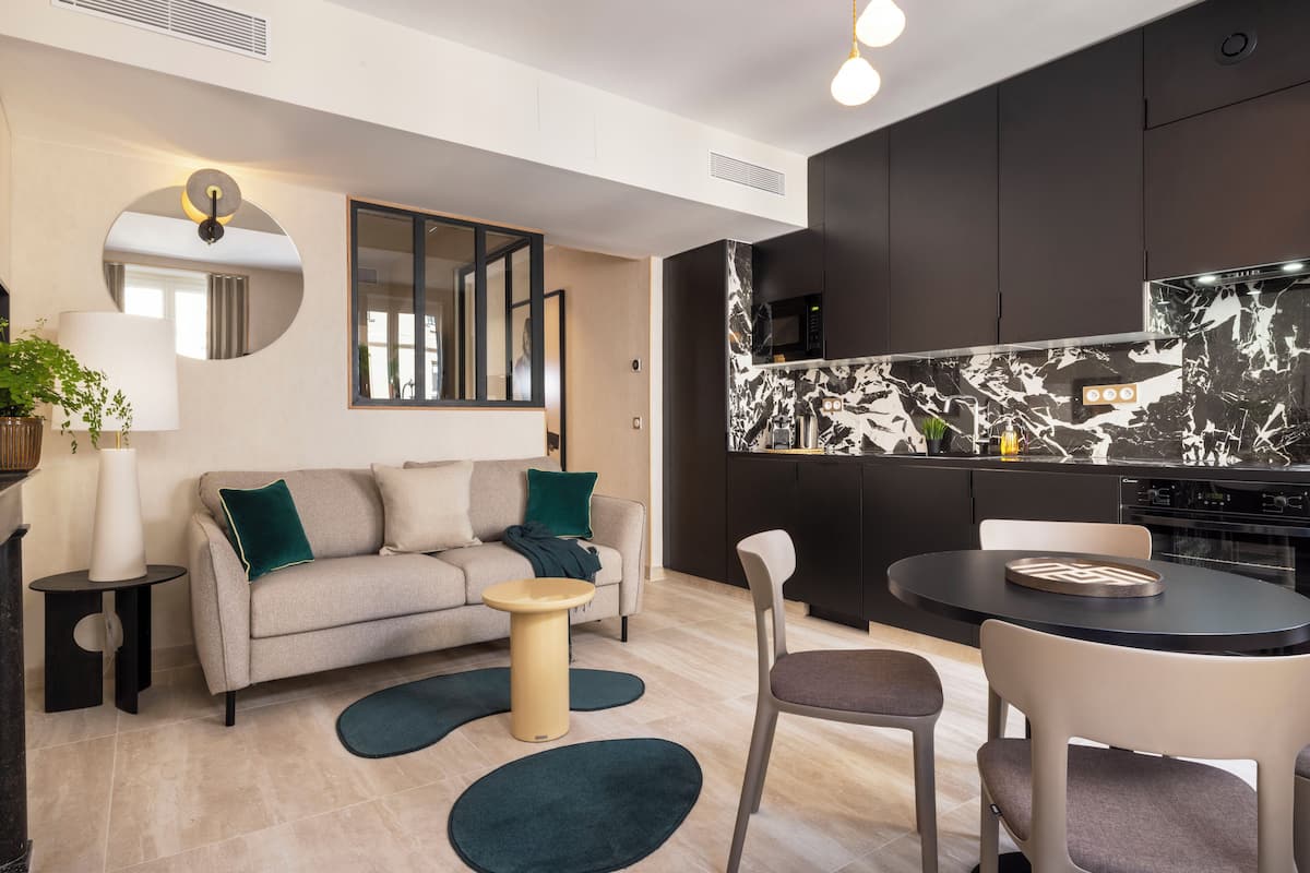 A modern living room with a sofa, coffee table, and chairs, a kitchen with dark cabinets and a marble backsplash, and a round mirror on the wall.