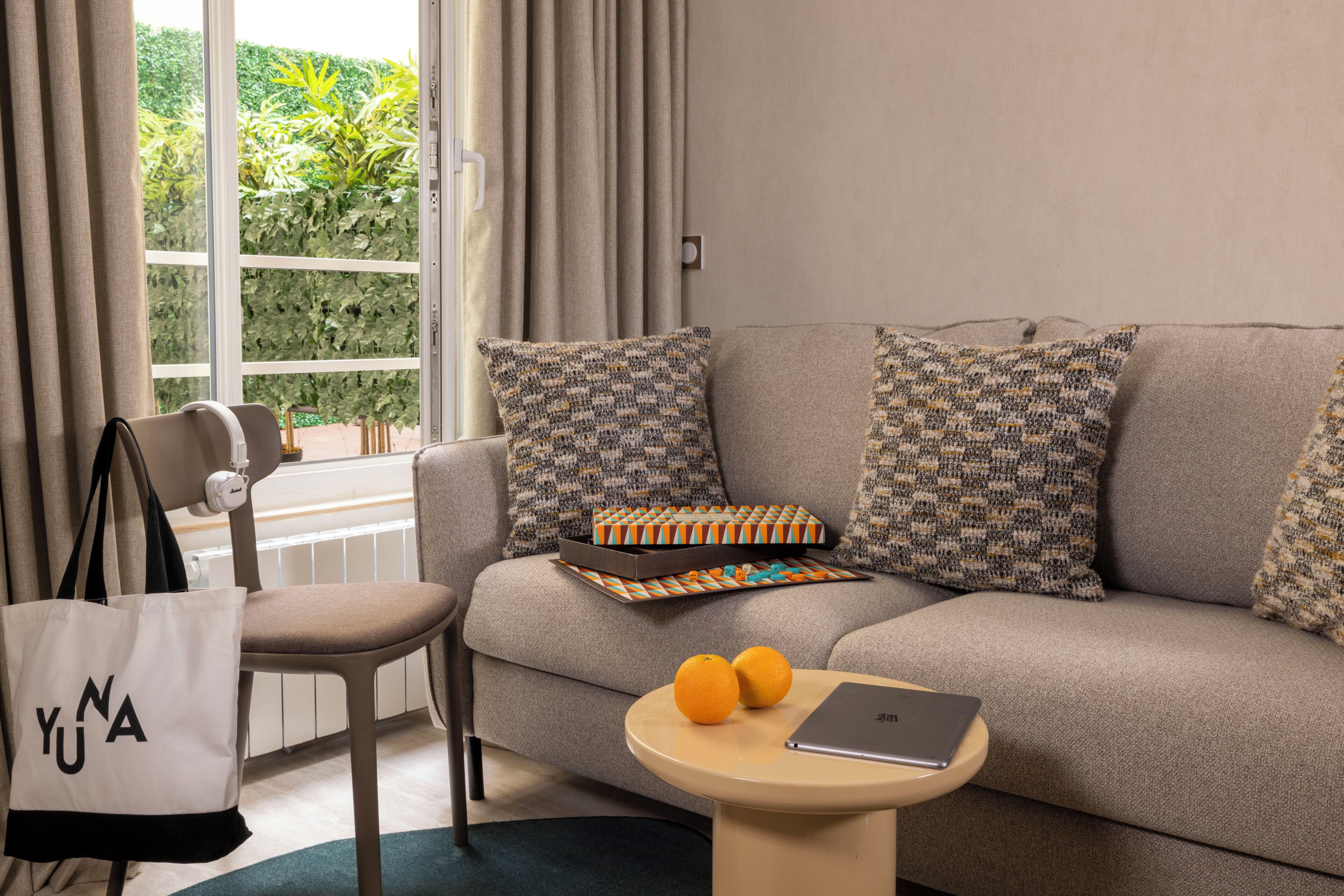 Una sala de estar moderna con un sofá, una butaca, una mesa con dos naranjas y una computadora portátil, y una ventana con cortinas.
