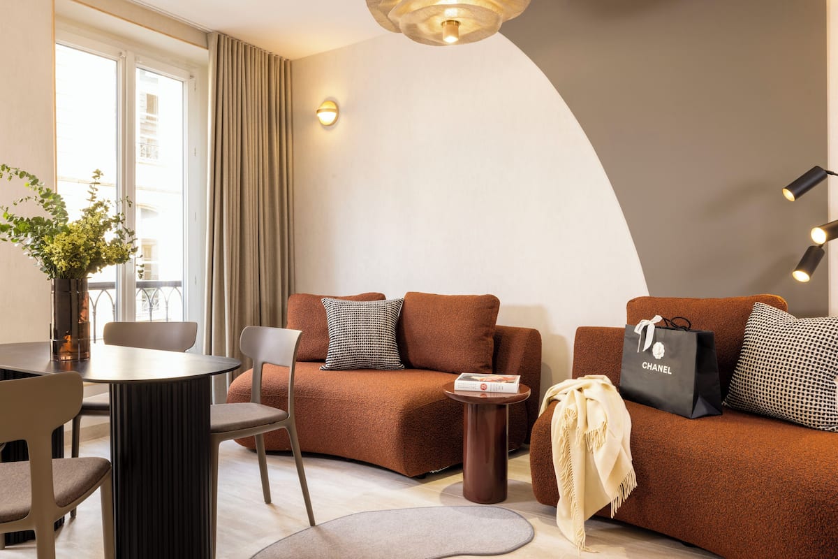 A modern living room with a brown sofa, a round table, and a chair. There is a vase with flowers on the table, and a Chanel shopping bag on the sofa.