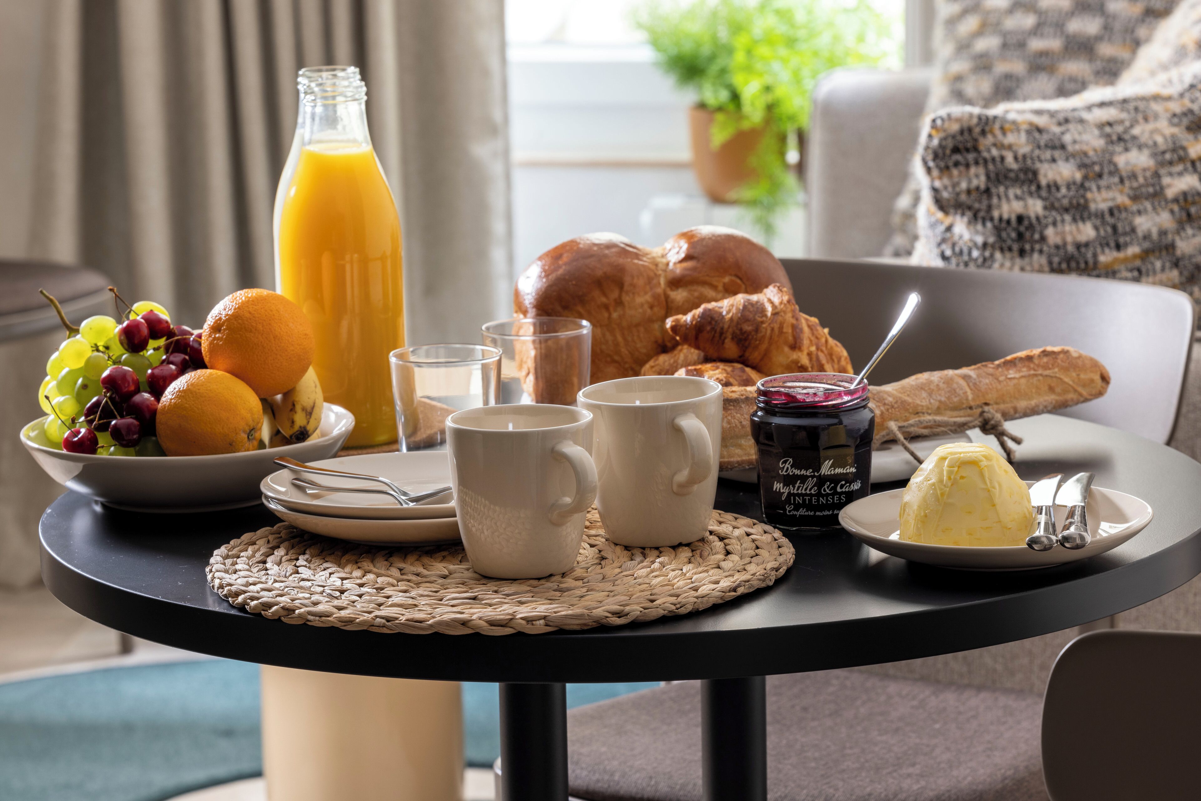 Une table de petit-déjeuner avec des croissants, une bouteille de jus d’orange, un saladier de fruits, deux tasses blanches et une assiette contenant un dessert.