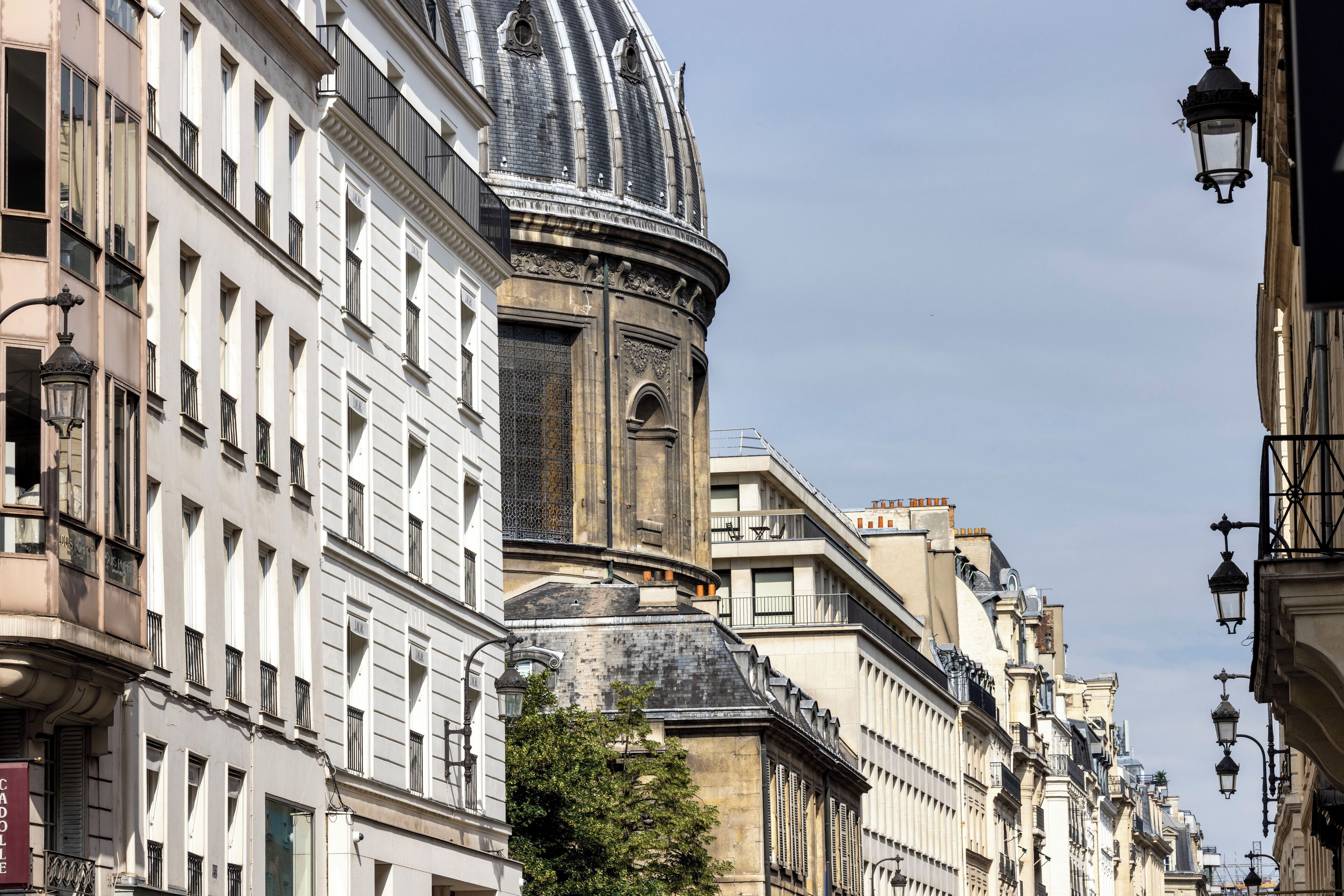 Vista da rua com edifícios históricos, incluindo um prédio com destaque e uma cúpula, e uma construção na esquina com topo arredondado.