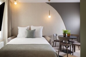 Moderni hotellihuone, jossa on suuri sänky, kaksi yöpöytävalaisinta, pieni pöytä tuoleineen ja ruukkukasvi.