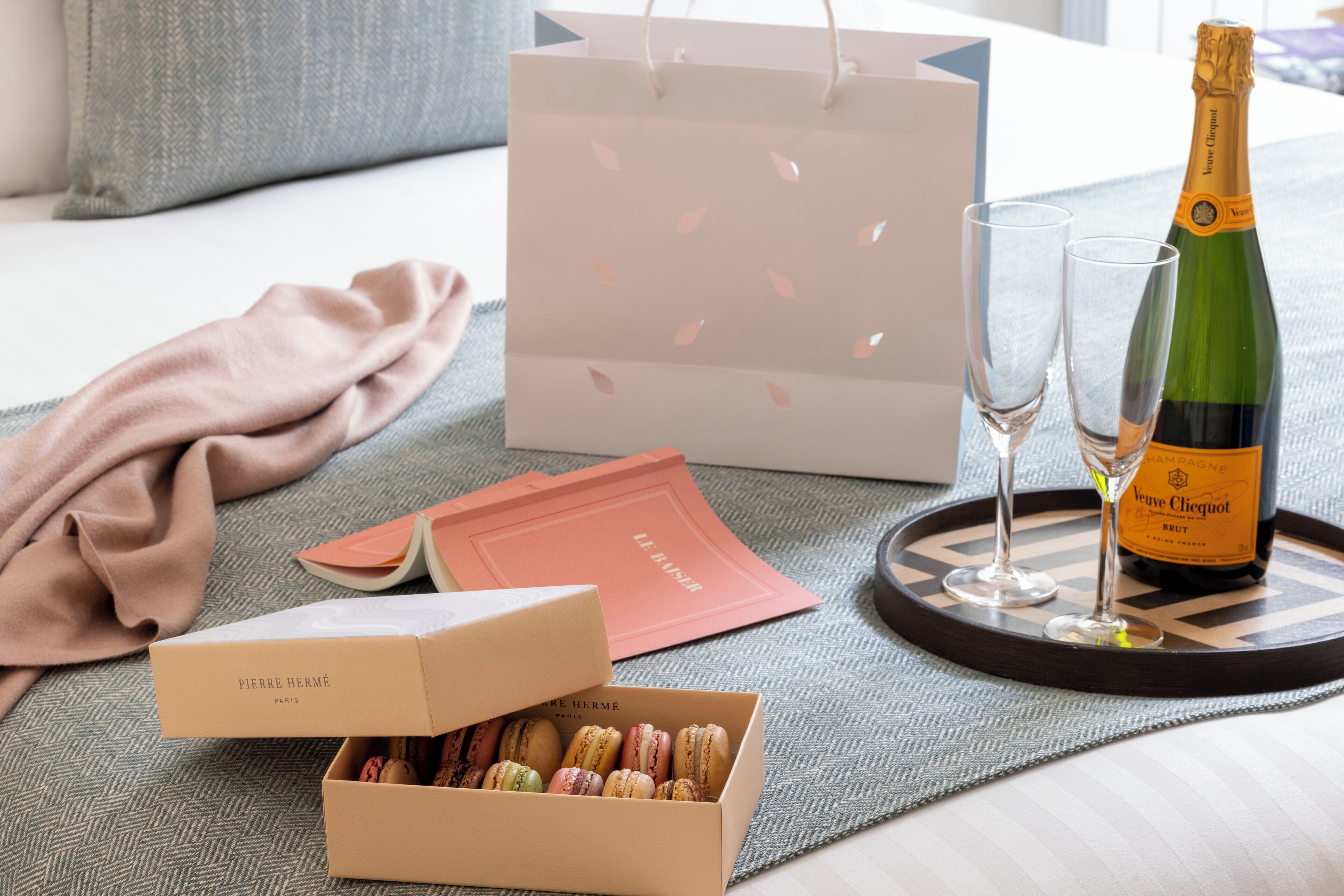 Una cama con una manta rosa, una caja de macarons, una botella de champán, dos copas y una bolsa de regalo sobre una bandeja.