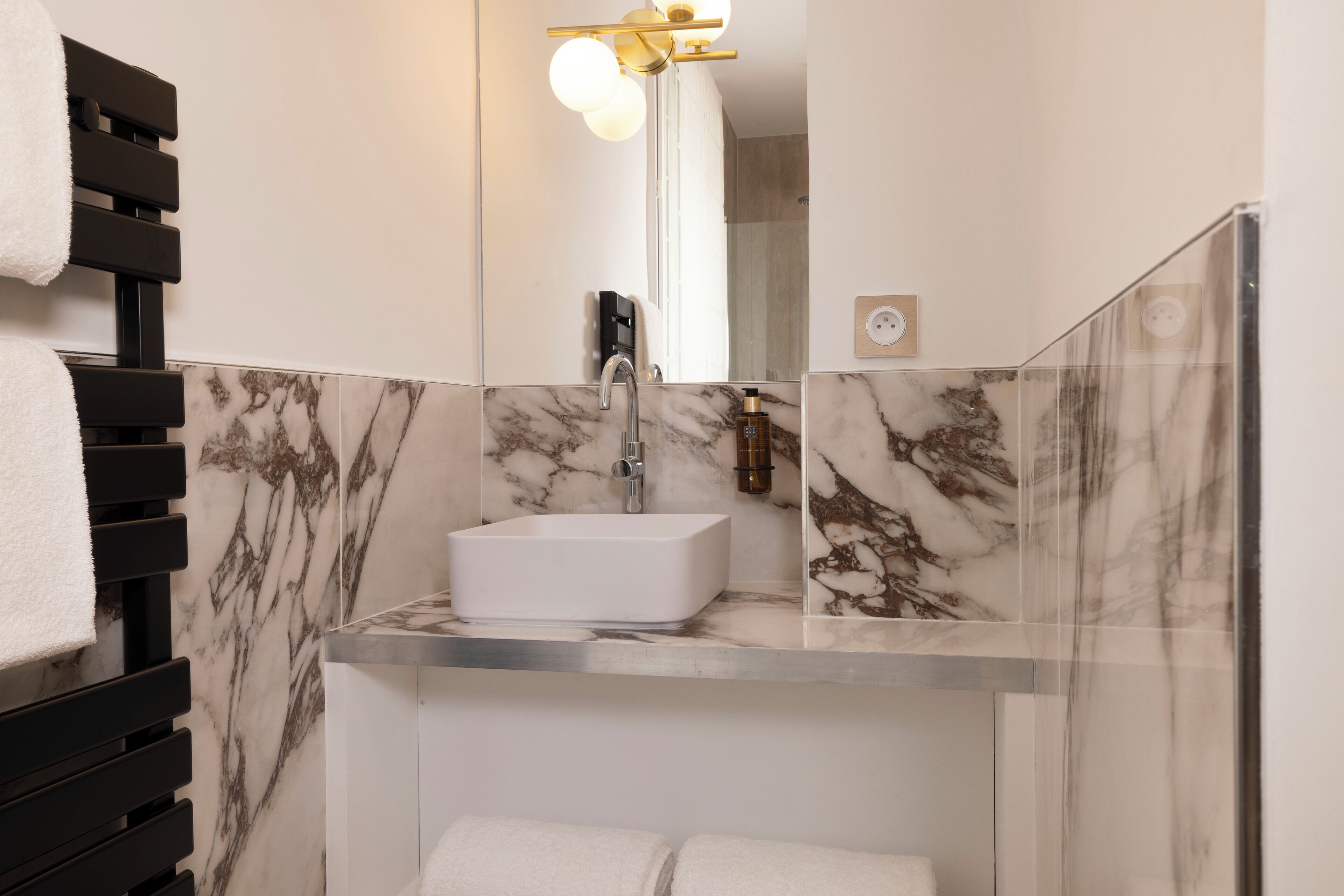 A modern bathroom with a white sink, marble-tiled walls, and a black towel rack.