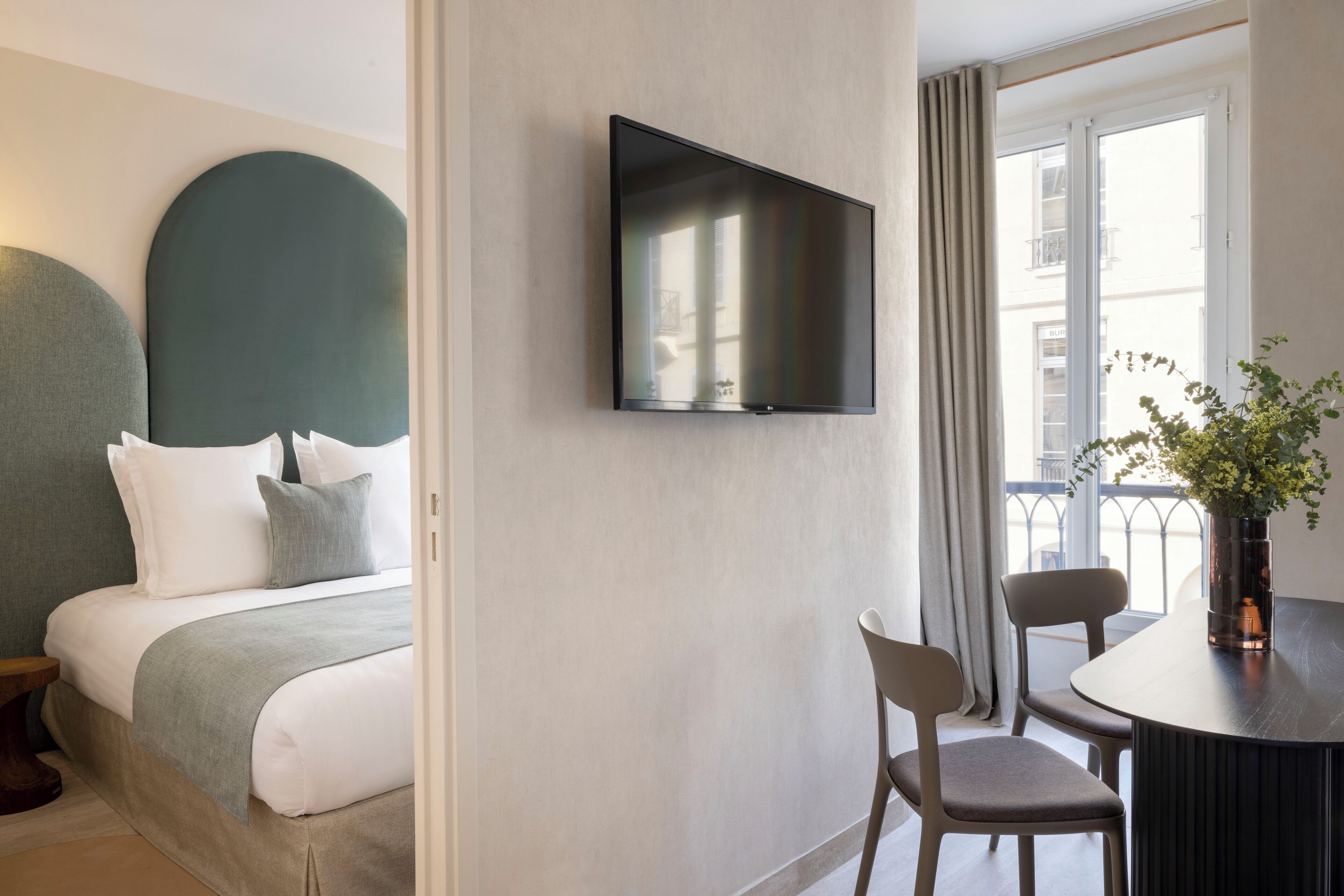 A modern hotel room with a bed, a flat-screen TV mounted on the wall, a dining table with chairs, and a window with curtains.