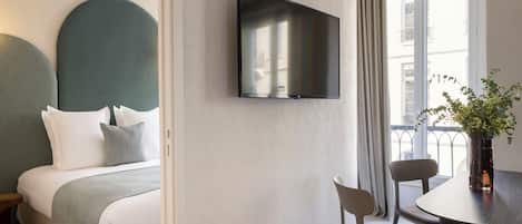 A modern hotel room with a bed, a flat-screen TV mounted on the wall, a dining table with chairs, and a window with curtains.